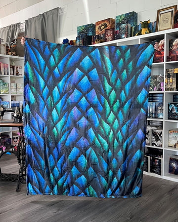 Das Produktbild zeigt eine Kuscheldecke mit blauen und grünen Drachenschuppen als Motiv.